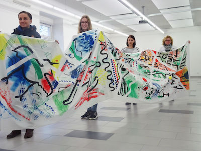 Vier Personen halten in einem hellen Raum ein langes, bunt abstrakt bemaltes Blatt hoch.