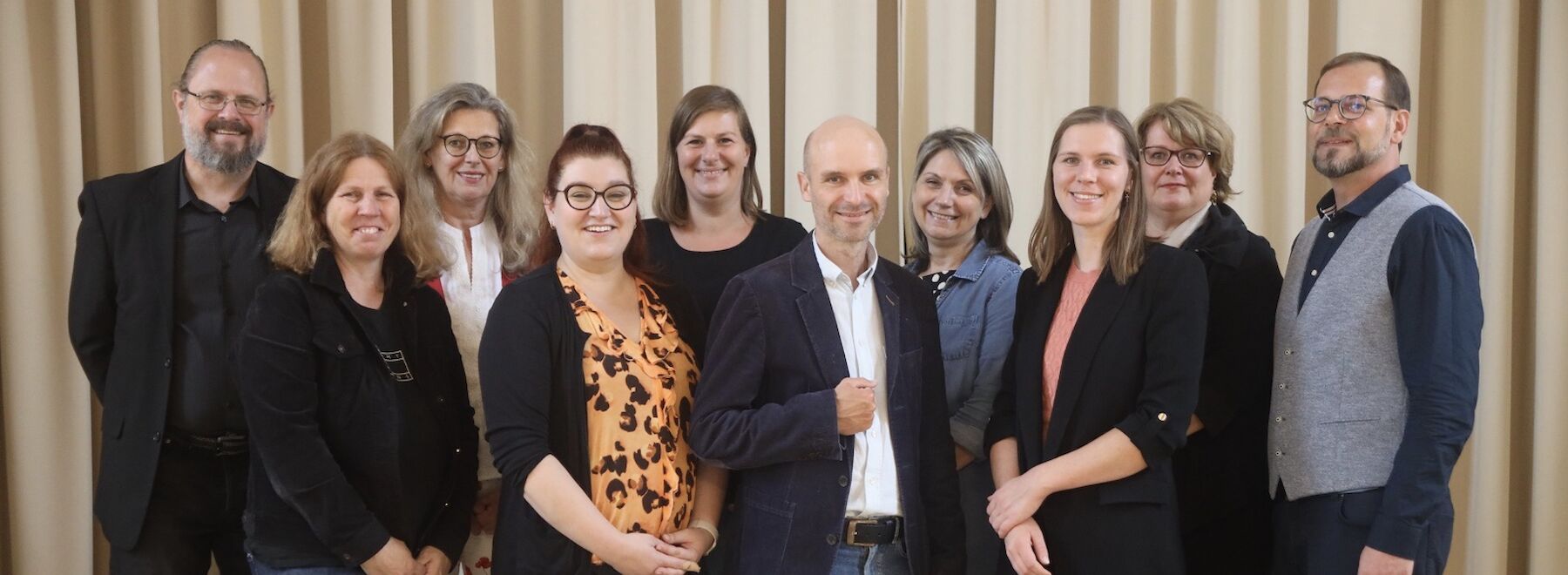 Elf Erwachsene stehen vor einer grünen Blätterwand und posieren für ein Gruppenfoto.
