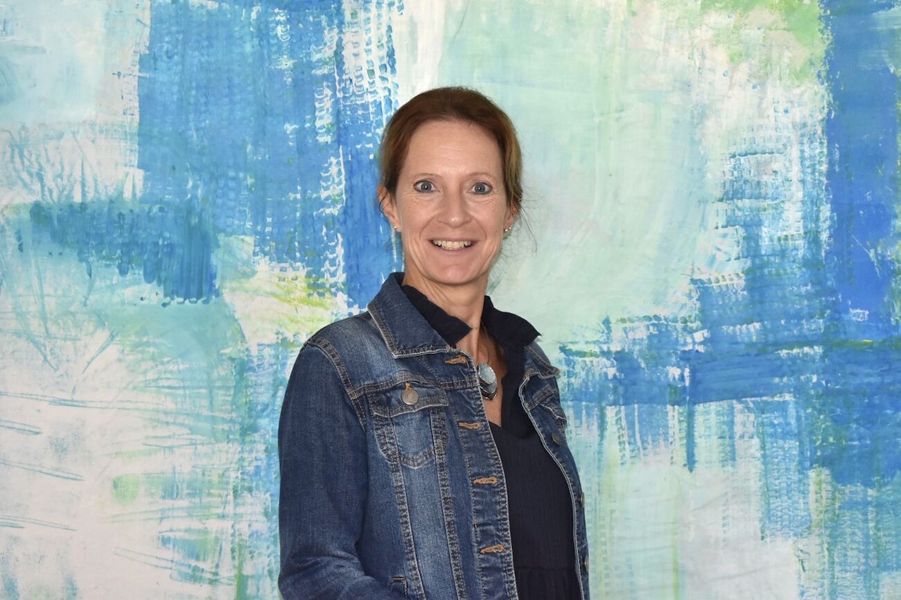 Lächelnde Frau mit grauem Haar in einer Jeansjacke, vor blauem Hintergrund stehend.
