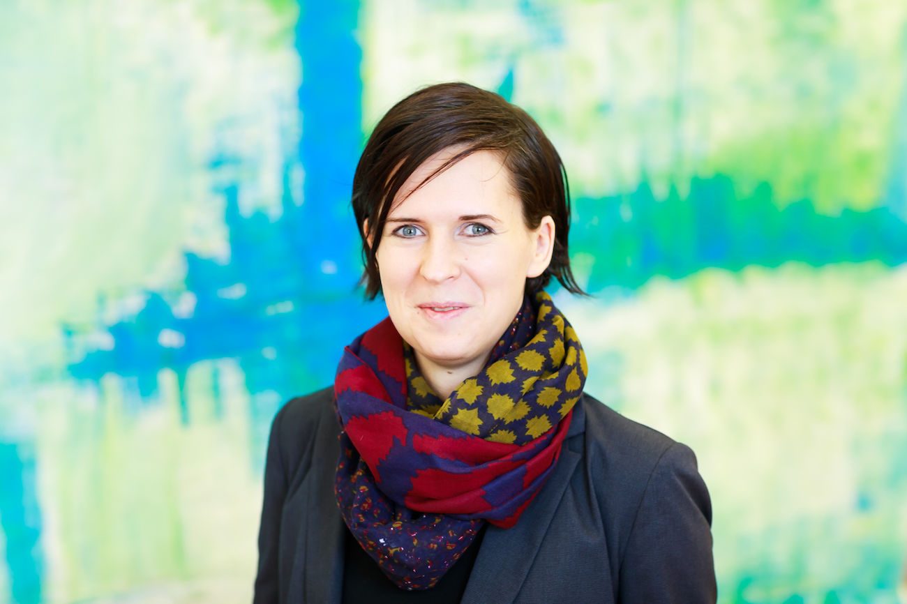 Frau mit kurzen dunklen Haaren, gemustertem Schal und Blazer, lächelt in die Kamera.