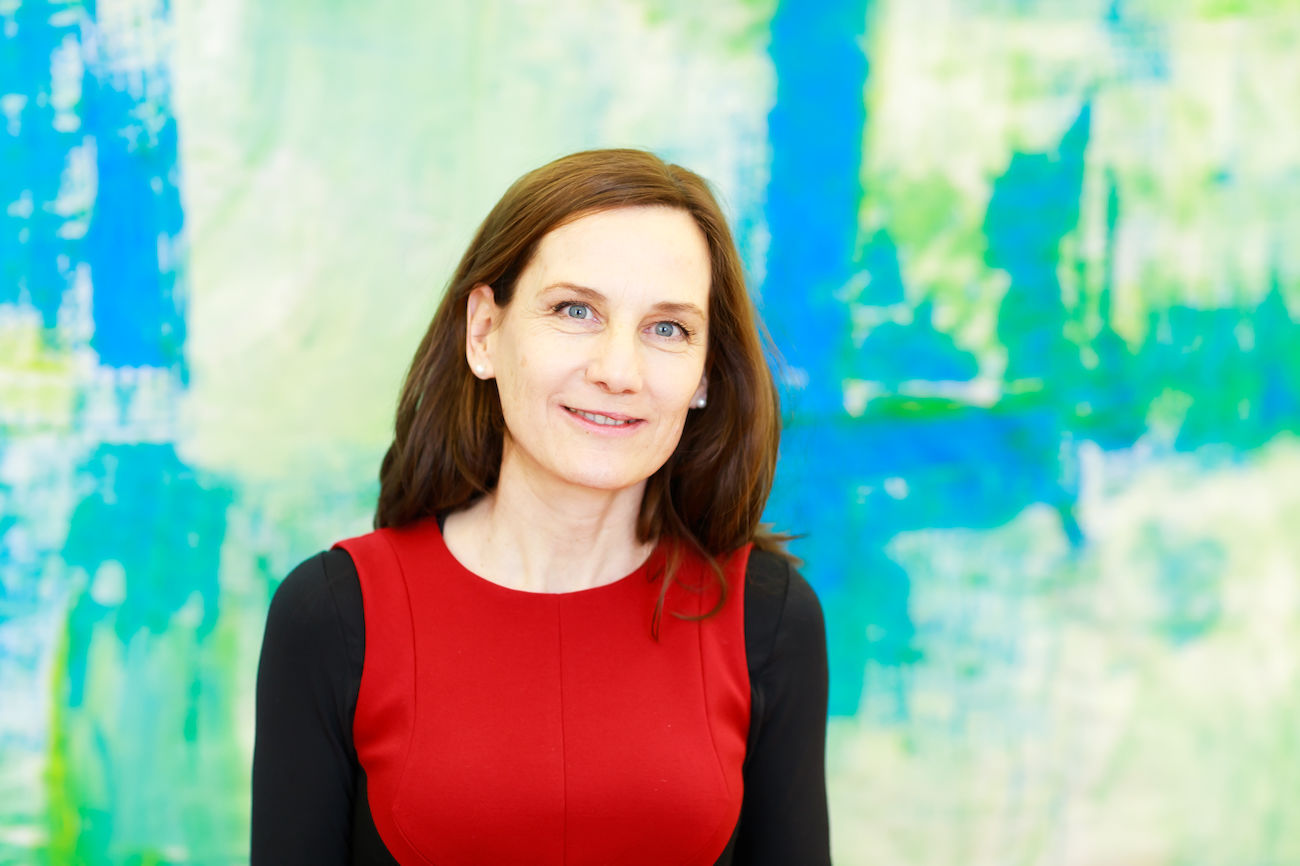 Frau mit braunen Haaren in einem roten Kleid steht vor einem blaugrünen Hintergrund.