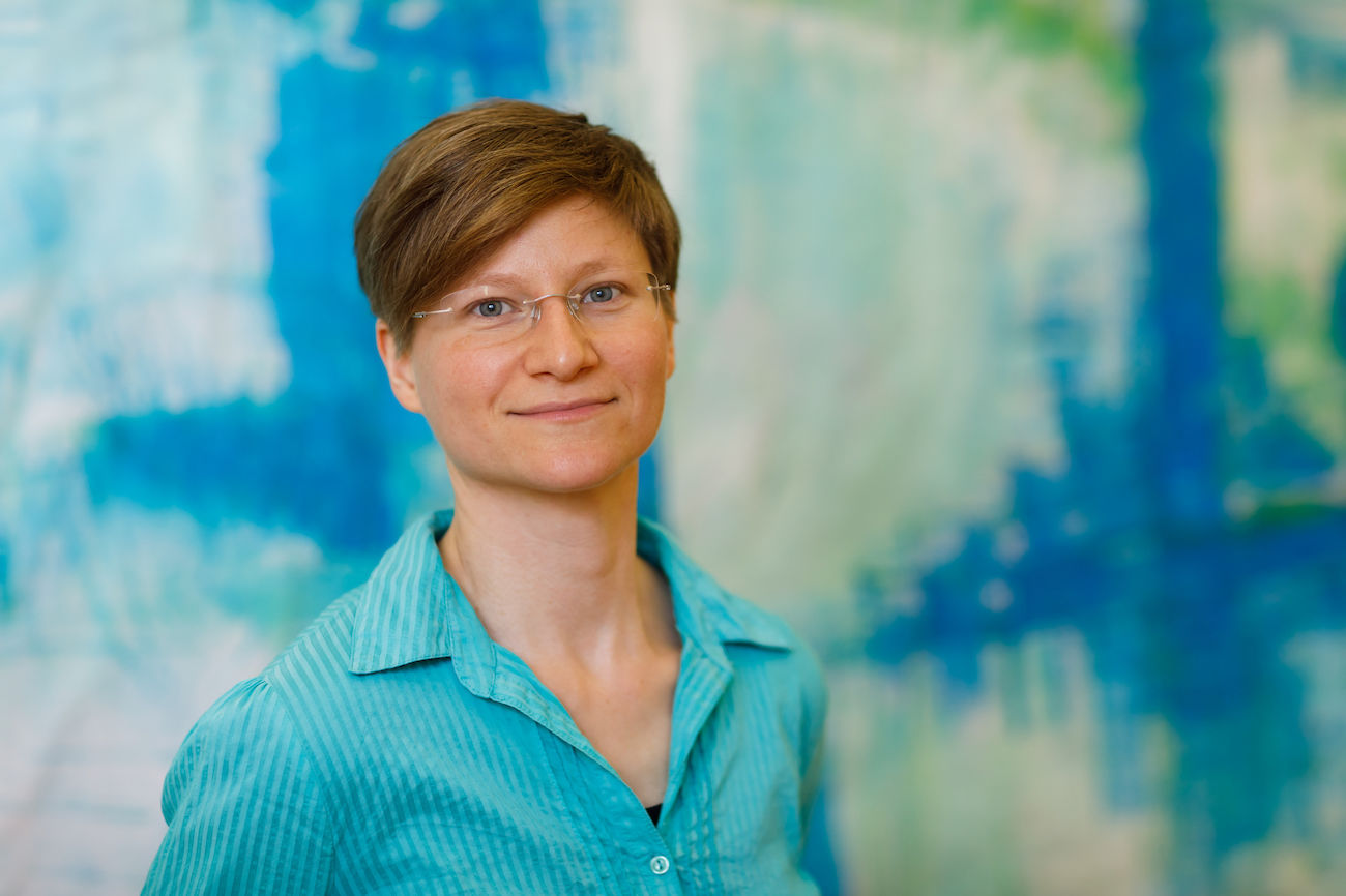 Person mit kurzen Haaren und Brille, die vor einem blauen abstrakten Hintergrund lächelt.