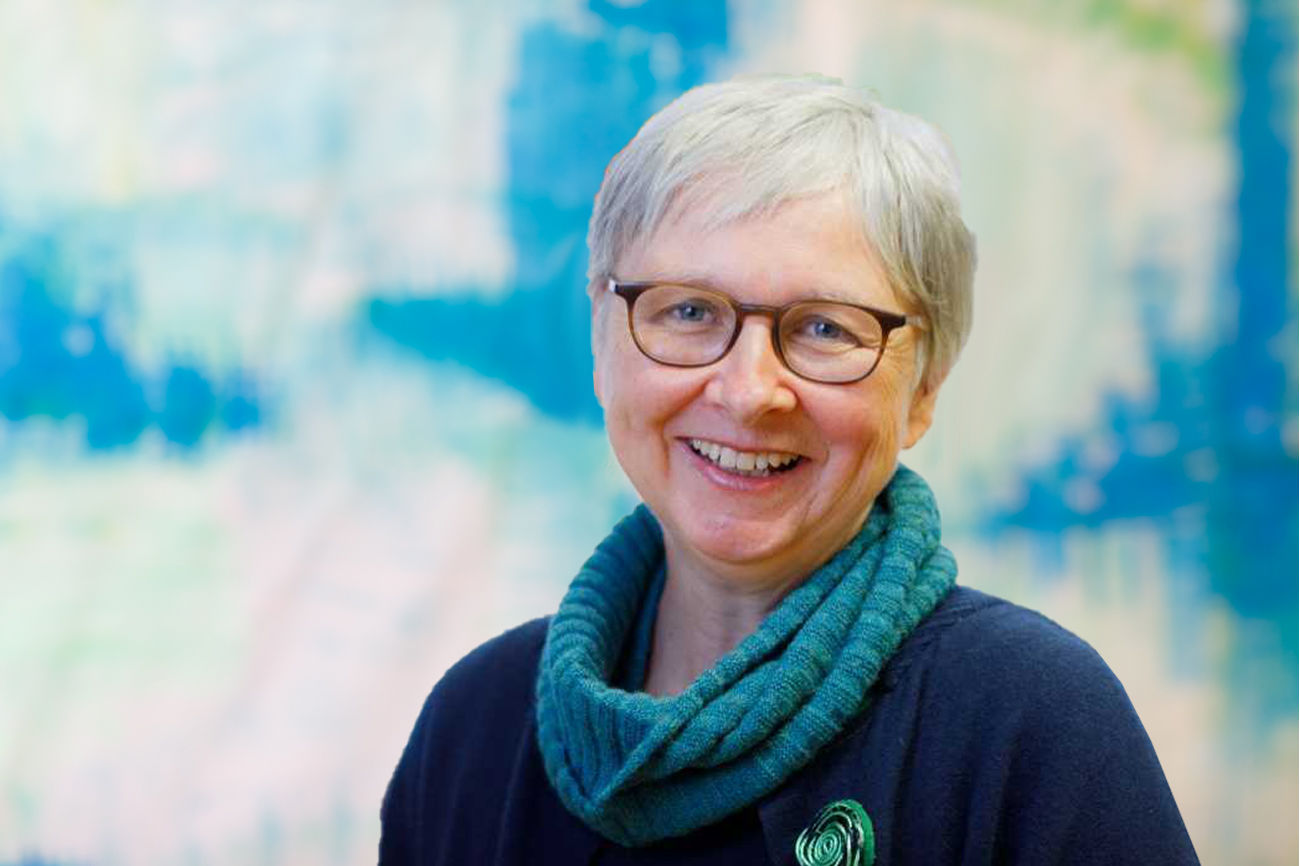 Lächelnde Frau mit kurzen grauen Haaren und Brille, trägt einen blaugrünen Pullover.