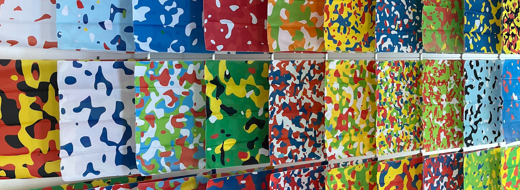 Bunte Gesichtsmasken mit Tarnmuster sind in Reihen an einer Wand ausgestellt.