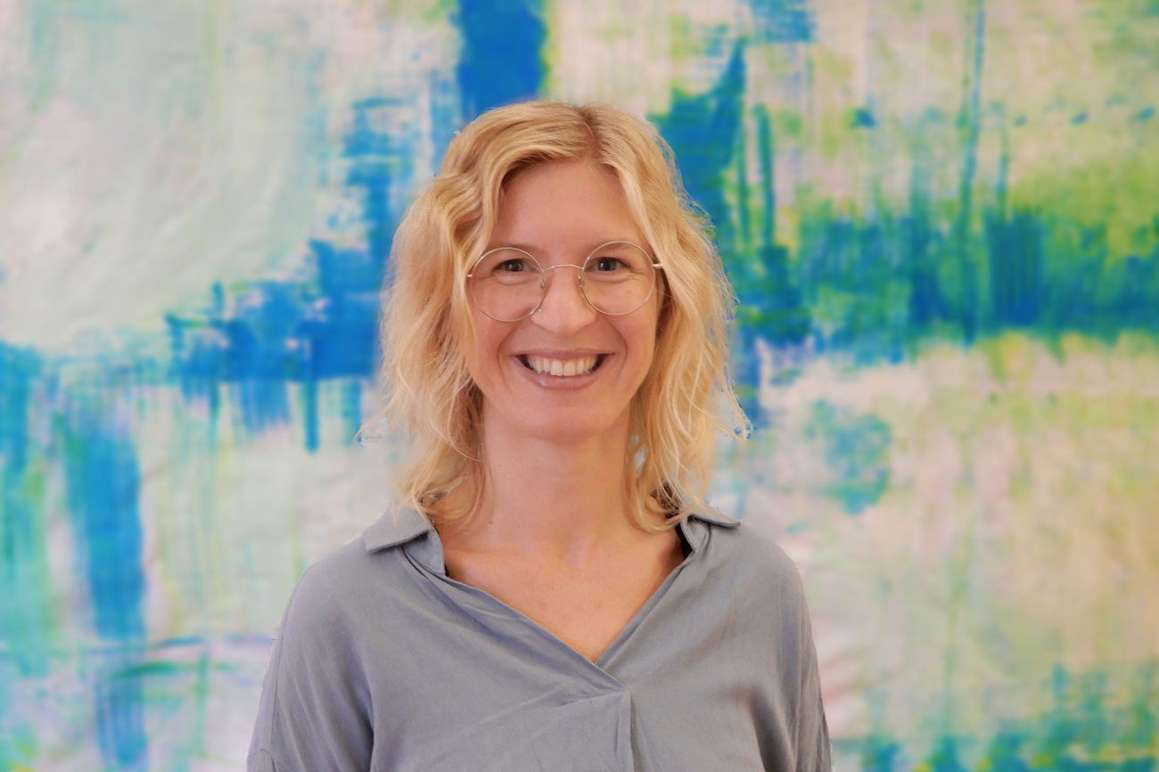 Lächelnde Frau mit blonden Haaren und Brille, die vor einem blauen abstrakten Hintergrund steht.