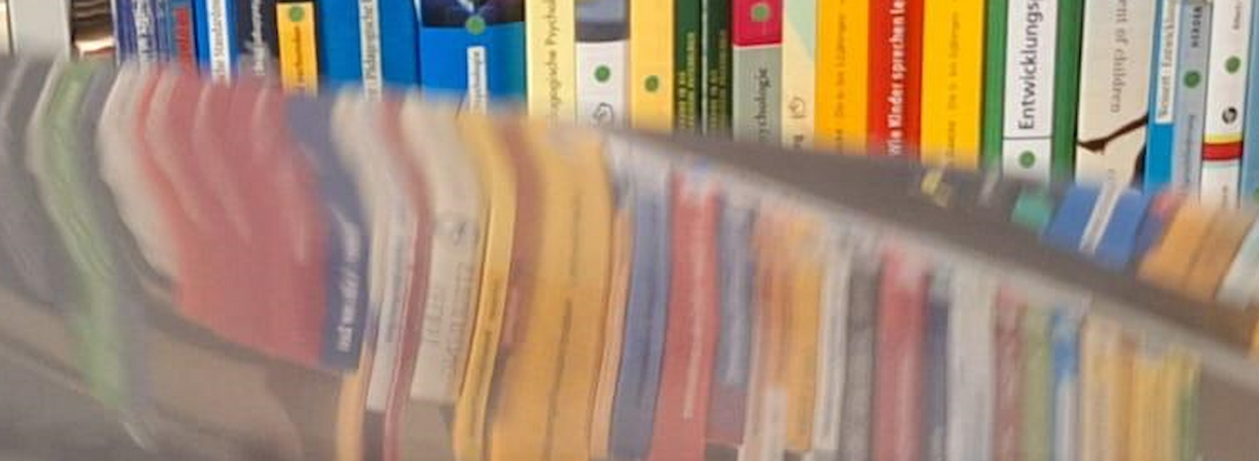 Unscharfes Bild von bunten Büchern auf einem Regal, teilweise durch ein durchsichtiges Blatt verdeckt.