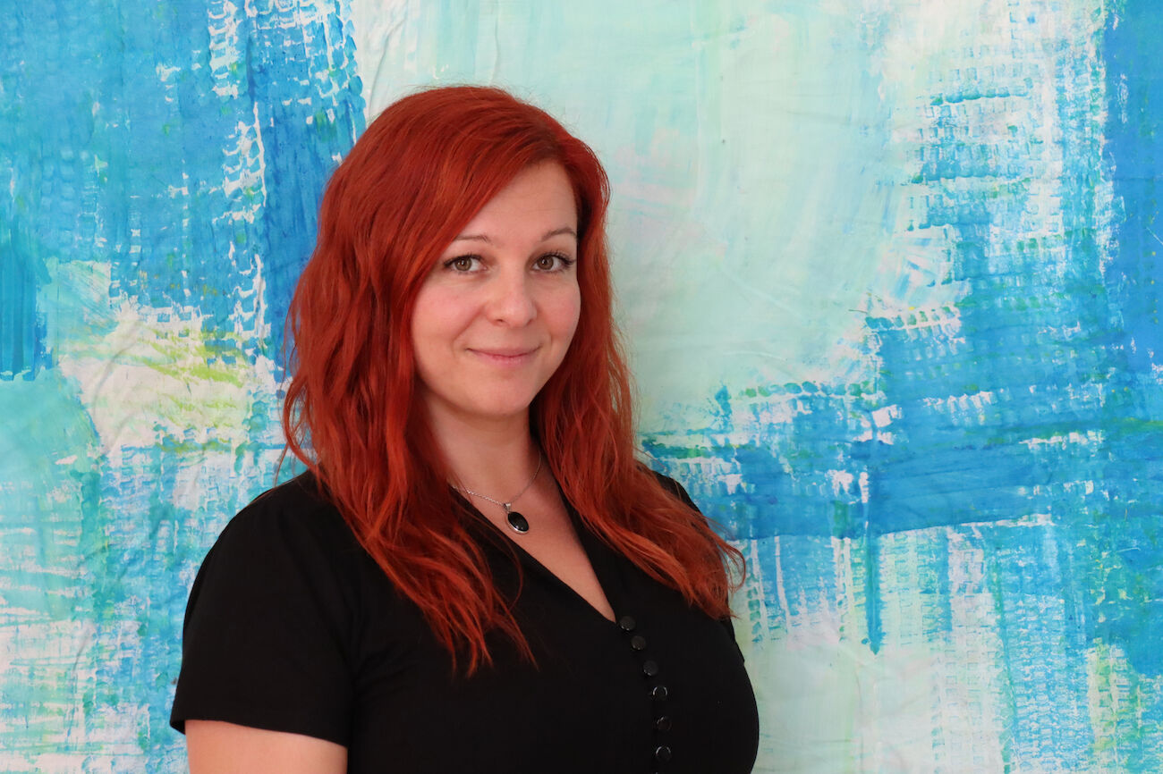 Frau mit langen roten Haaren in schwarzem Oberteil steht vor einem blauen abstrakten Hintergrund.