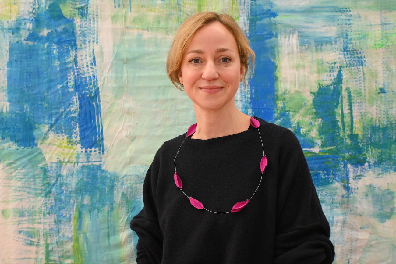 Lächelnde Frau mit blonden Haaren steht im Freien vor einem Holzzaun.