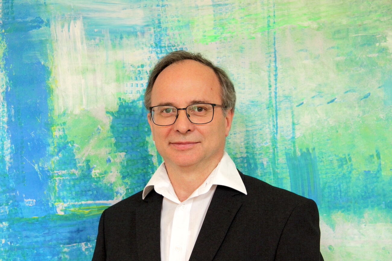 Mann mit Brille und Anzug steht vor einem blau-grünen abstrakten Hintergrund.