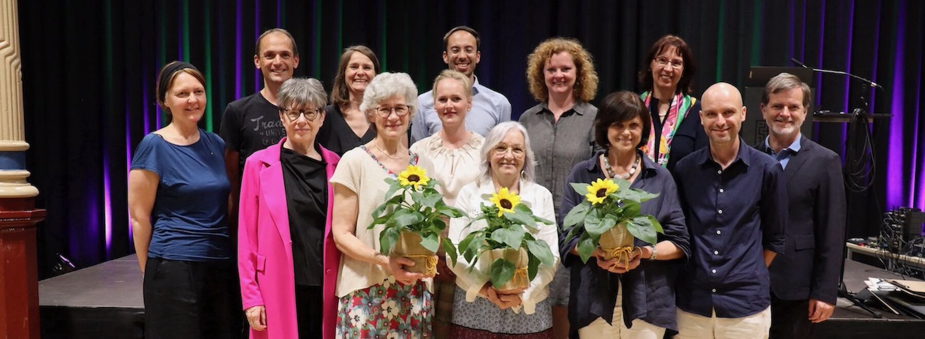 Eine Gruppe von Menschen posiert im Innenbereich; zwei Frauen davor halten Sonnenblumen in Töpfen.
