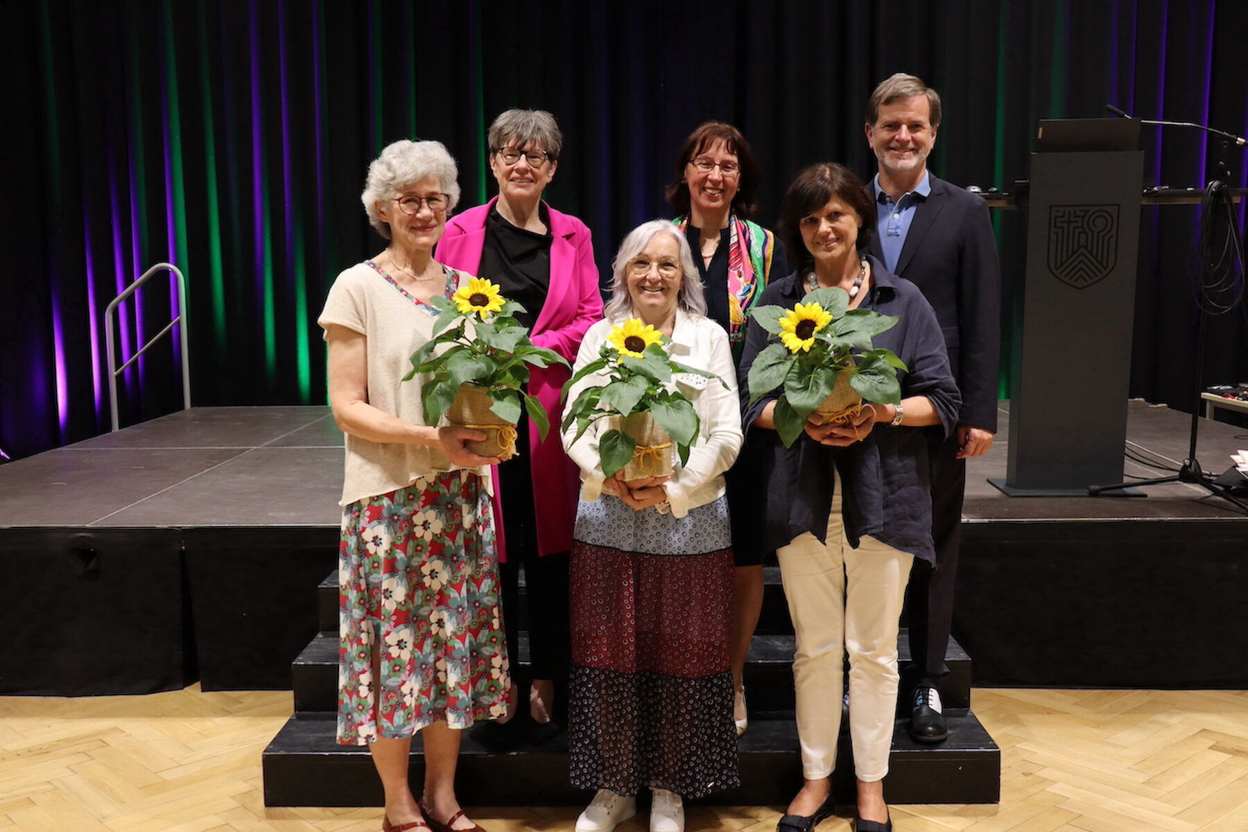 Fünf Frauen mit Sonnenblumen in der Hand und ein Mann stehen lächelnd zusammen auf einer Bühne.