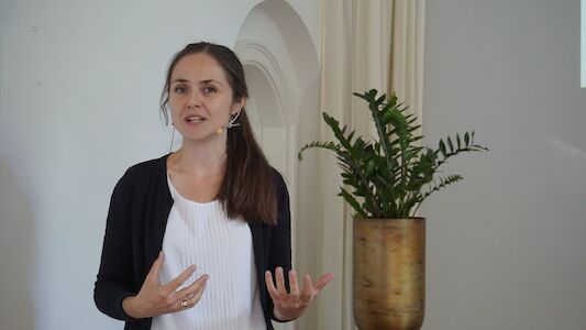 Frau spricht im Innenbereich neben einer grünen Topfpflanze und einer hellen Wand.