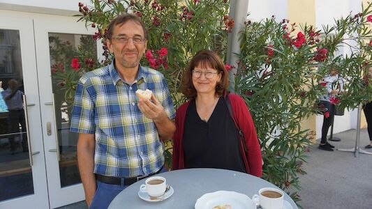 Zwei Personen genießen Kaffee und Gebäck an einem Tisch im Freien mit Blumen im Hintergrund.