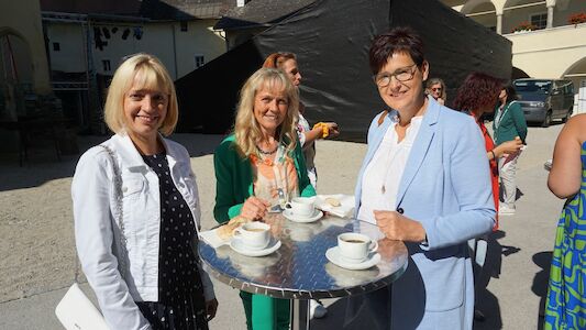 Drei Frauen stehen mit Kaffeetassen an einem runden Tisch und lächeln im Freien.