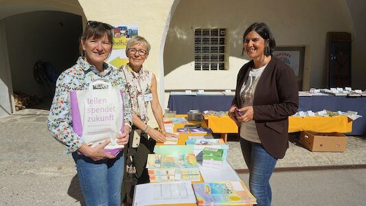 Drei Frauen stehen bei einer Veranstaltung im Freien an einem Tisch mit Büchern und Broschüren.