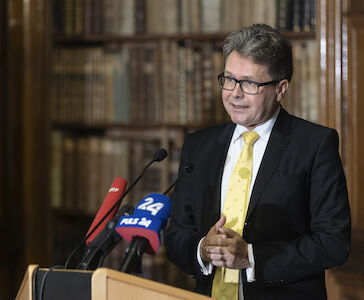 Mann in Anzug und gelber Krawatte spricht an einem Podium mit Mikrofonen in einer Bibliothek.
