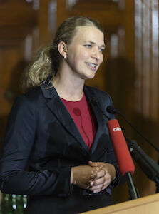 Frau im dunklen Blazer spricht an einem Podium mit Mikrofonen vor ihr.