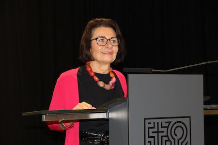Frau mit Brille und rosa Strickjacke spricht an einem Podium mit einem Logo.