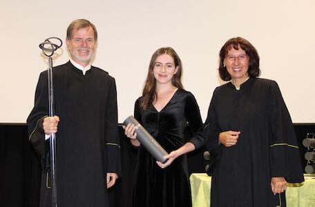 Drei Personen in schwarzen Roben bei einer Zeremonie, eine davon hält ein diplomähnliches Objekt.
