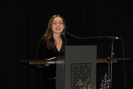 Frau spricht an einem Podium mit einem Mikrofon vor schwarzem Hintergrund.