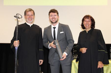 Drei Personen in Abendgarderobe, eine davon mit einem Diplom in der Hand, lächeln bei einer Veranstaltung.