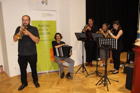Fünf Musiker spielen Blechblas- und Akkordeoninstrumente in einem kleinen Innenraum.