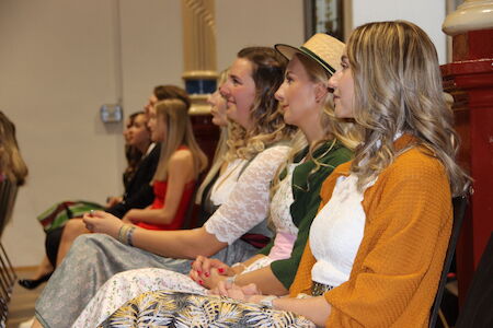 Mehrere Frauen in traditioneller Kleidung sitzen in einer Reihe und verfolgen aufmerksam ein Geschehen.