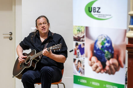 Mann spielt Akustikgitarre neben einem UBZ-Umweltplakat im Innenbereich.
