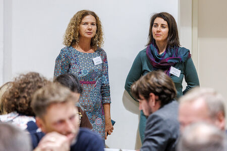Zwei Frauen stehen bei einer belebten Veranstaltung oder Besprechung in einem geschlossenen Raum vor einer weißen Wand.