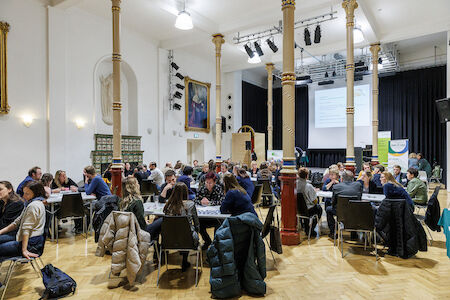 Personen, die während einer Gruppenveranstaltung oder eines Workshops an runden Tischen in einem großen Saal sitzen.