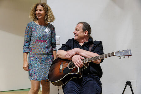 Eine lächelnde Frau steht neben einem sitzenden Mann, der eine Akustikgitarre hält.