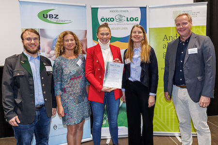 Fünf Personen stehen lächelnd zusammen, im Hintergrund sind Transparente und eine Urkunde zu sehen.