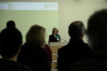 Eine Frau steht an einem Podium und spricht zu einem Publikum in einem schwach beleuchteten Raum.