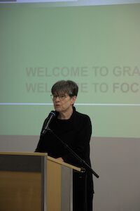 Frau spricht an einem Podium mit Mikrofon, hinter ihr wird Text projiziert.
