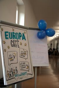 Plakat für das Europa Café neben blauen Luftballons und einem handgeschriebenen Willkommensschild.