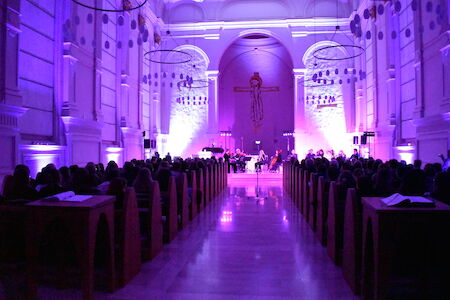 Der Kircheninnenraum ist während einer musikalischen Darbietung mit violettem Licht beleuchtet, das Publikum sitzt.