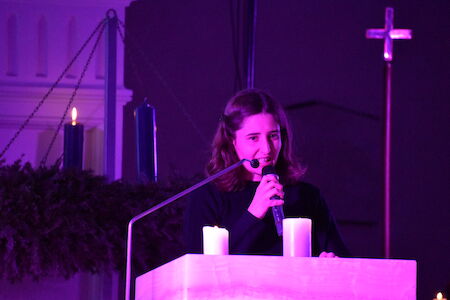 Eine Frau spricht in ein Mikrofon an einem Podium mit Kerzen, die in violettes Licht getaucht sind.