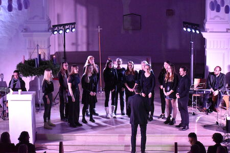 Ein Chor aus Frauen in Schwarz singt mit einem Dirigenten in einem violett beleuchteten Raum auf der Bühne.