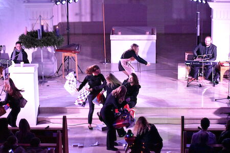 Eine Gruppe von Künstlern in schwarzer Kleidung tanzt auf einer Kirchenbühne, in der Nähe sind Musiker.