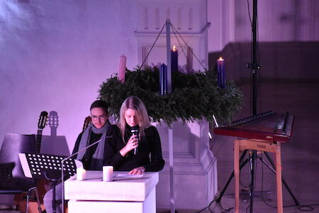 Zwei Personen sitzen in einem schwach beleuchteten Raum neben einem Adventskranz mit brennenden Kerzen.