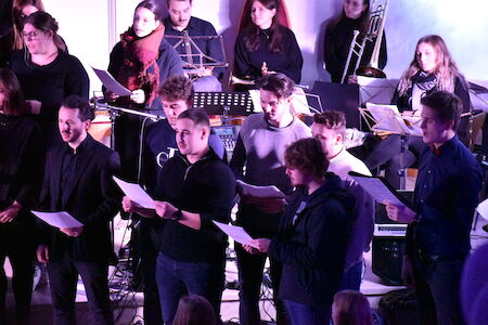 Eine Gruppe von Menschen singt in einem Chor, hinter ihnen spielen Musiker Instrumente.