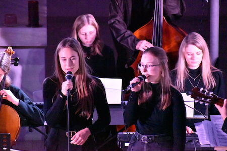 Zwei junge Frauen singen mit Mikrofonen vor einer kleinen Band auf der Bühne.