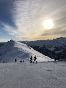 Silhouette der Skifahrer auf einem schneebedeckten Berghang bei Sonnenuntergang mit Gipfeln in der Ferne.