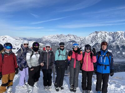 Sieben Personen in Skiausrüstung stehen zusammen auf einem schneebedeckten Berg, hinter ihnen liegen Gipfel.
