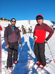 Zwei Männer in Skiausrüstung stehen unter einem klaren blauen Himmel auf einem schneebedeckten Hang.