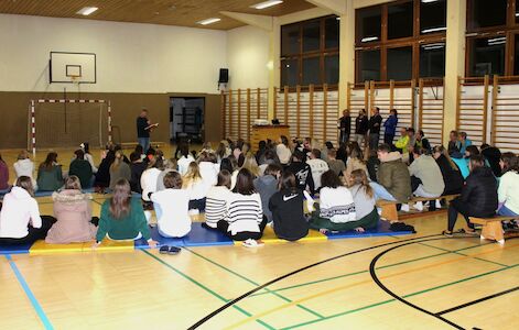Eine Gruppe von Menschen sitzt in einer Turnhalle auf Matten und blickt einem Redner zu.