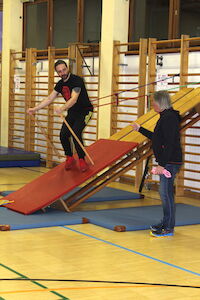 Mann balanciert auf einer schrägen Gymnastikmatte, während eine Frau in der Nähe steht und ein Seil hält.