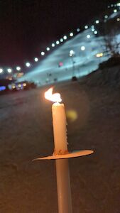Eine brennende Kerze, die nachts im Freien in der Nähe einer hell erleuchteten Skipiste gehalten wird.