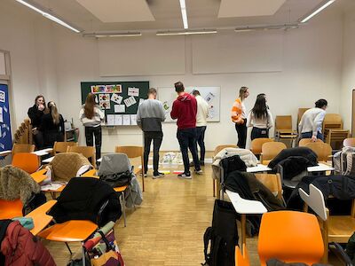 Schüler stehen und unterhalten sich in einem Klassenzimmer mit leeren Tischen und Papieren an der Wand.