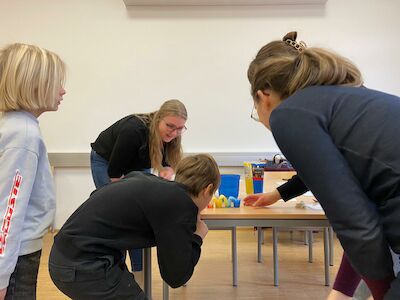 Vier Kinder und eine Frau beugen sich über einen Tisch und betrachten bunte Gegenstände.